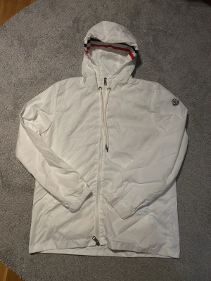Vit vindjacka från Moncler med huva - Säljer en fräsch vit vindjacka från Moncler med huva och dragkedja. Jackan har Moncler-logga på ärmen och coola ränder i rött, blått och svart på insidan av huvan. Långärmad och tillverkad i polyester, perfekt för blåsiga dagar.