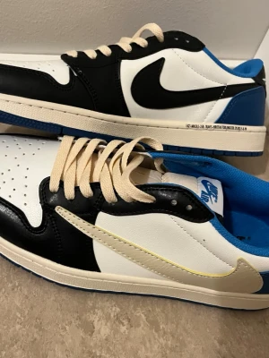 Nike Air Jordan 1 Low Travis Fragment - Nike Air Jordan 1 Low Travis Scott x Fragment med svart, vit och blå färg. Skorna har omvänd Swoosh i vitt, klassisk snörning och detaljer som broderad logga och text på hälen. Tillverkade i skinn med blå yttersula och perforerad tå för extra stil.
