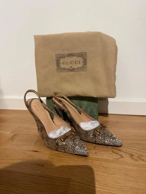 Gucci klackar - Säljer ett par lyxiga Gucci klackar med spetsig tå, brun färg och glitter stenar. Klackarna har metalldetaljer med öppen häl. Materialet är skinn och textil, och klacken är medelhög 8cm. Kommer med kartong och dustbag. Passar även perfekt till student 