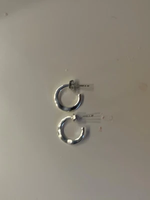 Örhängen  - Bilden visar ett par silvriga örhängen i form av små hoops. Ingen väst finns på bilden. Dom är platta
