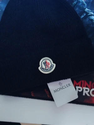 Svart Moncler mössa i ull - Snygg svart mössa från Moncler med klassisk logga framtill. Tillverkad i mjuk ull som håller dig varm och stilren under kyliga dagar. Perfekt accessoar för dig som vill ha både värme och streetstyle.