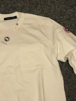 Vit långärmad tröja från Canada Goose - Snygg vit långärmad tröja från Canada Goose med klassisk rund hals och en praktisk bröstficka. Tröjan har en diskret logotyp på ärmen och är gjord i mjuk bomull som känns skön mot huden. Perfekt för dig som gillar stilrena och enkla plagg.