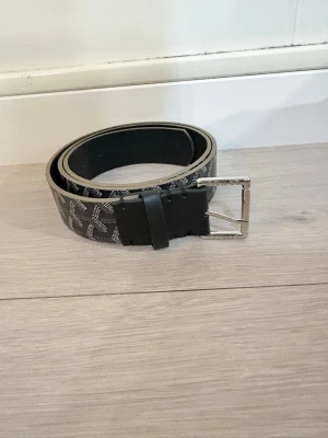 Svart bälte med mönster och silverfärgat spänne - Snyggt svart bälte med ett stilrent mönster i vitt och silverfärgat metallspänne. Bältet har en klassisk form och passar perfekt till jeans eller kostymbyxor. Materialet känns slitstarkt och har en lyxig känsla.