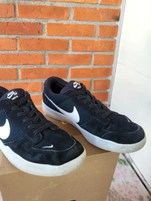 Nike SB Force 58 svarta sneakers 42 - Nike SB Force 58 sneakers i svart med vit swoosh och vit sula. Skorna har snörning och är låga med rund tå. Ovandelen är i mocka och textil, vilket ger en skön och stilren vibe. Perfekta för dig som gillar streetwear och skate.