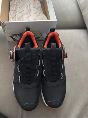 Solid Gear revolution 2  - Svarta sneakers från Solid Gear med orange detaljer och vit sula. Skorna har BOA-snörning för enkel justering och en platt, mönstrad sula i orange. Insidan är fodrad med orange mesh och ovandelen är tillverkad i syntetmaterial. Perfekt för dig som gillar sportig stil och smarta funktioner. Se sista bild för lite information och en beskrivning om skorna.