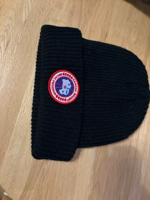 Canada Goose mössa - Säljer en svart ribbstickad mössa från Canada Goose med klassisk rund logotyp framtill i rött, vitt och blått. Aldrig använd ❤️