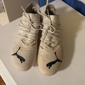 Vita fotbollsskor Puma x Liberty of London  - Snygga fotbollsskor från Puma i ljusbeige med svarta och röda loggor. Det är kvinno fotbollsskor. Skorna har blommigt mönster bak, snörning och plös i stickad syntet. Sulan är vit med orange dobbar för bra grepp på planen. Perfekta för dig som vill sticka ut på matchen! 