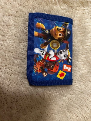 Blå Paw Patrol plånbok - Säljer en färgglad Paw Patrol plånbok med blått tyg och tryck av flera karaktärer från serien. Plånboken har detaljer i olika nyanser av blått och är tillverkad i syntetmaterial. Perfekt för att hålla koll på kort och småpengar. 55 kr/st 