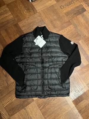 Svart quiltad pufferjacka Moncler - Svart pufferjacka från Moncler med quiltad framsida och stickade ärmar. Jackan har dragkedja, ribbad krage och Moncler-logga på ärmen med NFC scan. Insidan har ett coolt serietryck och märkestaggar. Perfekt för dig som vill ha en snygg och modern look. Oanvänd.
