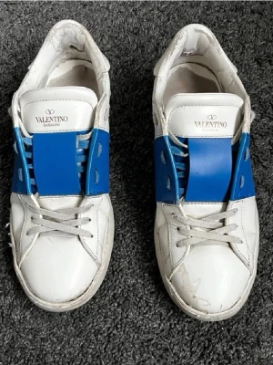 Valentino Garavani sneakers med blå detalj - Säljer ett par snygga Valentino Garavani sneakers i vitt skinn med bred blå panel över snörningen. Skorna har rund tå, platt sula och klassisk snörning. Perfekt för dig som gillar exklusiva märken och vill sticka ut med en cool färgdetalj.