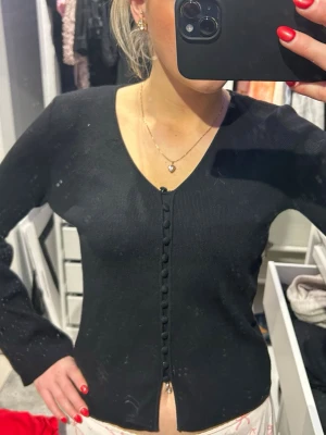 Svart kofta från Veromoda - Säljer en svart, ribbad kofta från AWARE i storlek XS. Koftan har v-ringning och dekorativa knappar längs hela framsidan. Materialet är mjukt och stickat, perfekt för lager-på-lager. Långärmad och stilren design som funkar till många outfits.