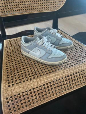Nike Dunk Low i ljusgrå och vit - Säljer ett par Nike Dunk Low sneakers i ljusgrå och vit färg. Skorna har klassisk snörning, rund tå och platt sula. Perfekt för dig som gillar stilrena och sportiga sneakers med ikonisk design från Nike.