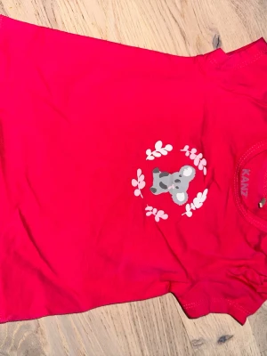 Röd t-shirt med koala från Kanz Kanz röd t-shirt, storlek 68 6M - Säljer en röd t-shirt från Kanz i storlek 68 med ett gulligt tryck av en grå koala omgiven av vita blad på bröstet. T-shirten har korta ärmar och rund hals. Perfekt för dig som gillar söta och färgglada plagg.Söt röd t-shirt med en koala tryckt på framsidan. Den har korta puffärmar.