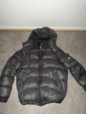 Svart Moncler pufferjacka med huva - Säljer en svart pufferjacka från Moncler med huva och dragkedja. Jackan har två fickor med dragkedja framtill och en Moncler-logga på ärmen. Insidan har ett unikt serietidningsmärke. Perfekt för kalla dagar och riktigt snygg streetstil.