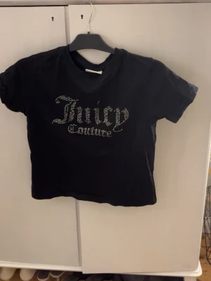 Svart Juicy Couture t-shirt med strass - Svart t-shirt från Juicy Couture med glittrande strassdetaljer på bröstet i logotypen. Modellen är kortärmad och har en klassisk rund hals. Materialet är mjuk bomull och passformen är normal. Perfekt för dig som gillar statement-plagg och ikoniska märken.