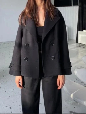 Svart kappa - Säljer en svart kort kappa med bred krage och dubbelknäppning framtill från Design By Si. Jackan har stora knappar och fickor på sidorna. Modellen är loose fit och har långärmade ärmar med knappdetaljer vid mudden. Perfekt för dig som gillar klassisk stil med en modern twist. Passar perfekt till höst och vår! Säljer då den inte kommer till använding💕