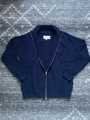 Marinblå stickad kofta från Maison Margiela - Säljer en marinblå stickad kofta från Maison Margiela med dubbel dragkedja framtill och ribbade muddar. Koftan har lång ärm och klassisk krage, perfekt för dig som gillar stilrena och tidlösa plagg. Diskreta vita stygn i nacken som signaturdetalj.