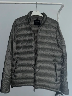 Grå pufferjacka från Peak Performance - Säljer en grå pufferjacka från Peak Performance med horisontella quiltade sektioner och två sidofickor med dragkedja. Jackan har ståkrage och hel dragkedja framtill. Perfekt för kyliga dagar och enkel att matcha med din stil.