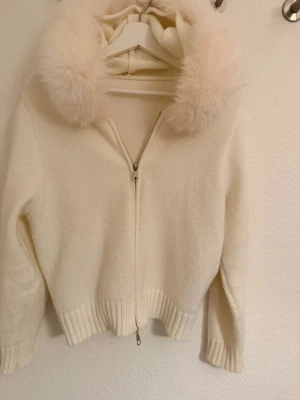 Vit stickad jacka med fluffig huva - Supermysig vit stickad jacka med dragkedja och fluffig pälsdetalj runt huvan. Jackan har ribbade muddar vid ärmslut och nederkant, och är långärmad. Perfekt för dig som vill ha en cozy och stylish look under kyliga dagar. Nästintill aldrig använd .