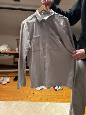 Oversize skjorta - Säljer en stilren ljusgrå skjorta med två stora bröstfickor och knappar framtill. Skjortan har klassisk krage och är tillverkad i ett mjukt material som känns skönt mot huden. 