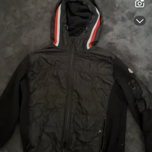 Svart vindjacka med huva och röd/vit detalj - Svart vindjacka från Moncler med huva som har röda och vita ränder. Jackan har dragkedja framtill, fickor med dragkedja och en patch på ärmen. Materialet är polyester och jackan har en quiltad front. Perfekt för dig som gillar sportig stil.