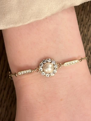 Armband från Lily & Rose - Snyggt armband i guldfärg med en rund pärla i mitten, omgiven av glittrande strass. Kedjan är tunn och har små detaljer med strass på sidorna. Perfekt för dig som gillar eleganta smycken med lite extra bling. 🤍