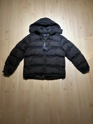 Svart pufferjacka från Polo Ralph Lauren- Storlek:M - Svart pufferjacka från Polo Ralph Lauren med huva och två sidofickor med dragkedja. Jackan har en röd broderad logga på bröstet och är tillverkad i polyester. Perfekt för kalla dagar och har en snygg, modern look. storlek är M