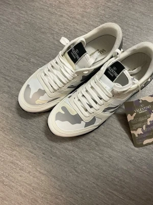 Valentino Garavani Rockrunner sneakers - Säljer ett par Valentino Garavani Rockrunner sneakers i ljusbeige och vitt med snyggt kamouflagemönster. Skorna har rund tå, snörning och en robust svart gummisula med mönstrad undersida. Materialet är en mix av skinn och textil, med coola detaljer på hälen.