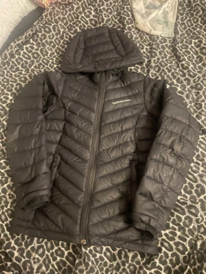 Svart dunjacka Peak Performance XS - Svart dunjacka från Peak Performance i storlek XS. Jackan har huva, dragkedja framtill och är quiltad med snygga diagonala sömmar. Perfekt för kyliga dagar och har en stilren look som funkar till det mesta. Materialet är syntet och dun. Det finns ett litet hål vid armen men de är lagat använd en vår sen sprillans ny