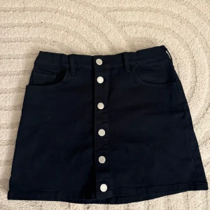 Svart kortkjol med knappar från GANT - Säljer en svart kortkjol från GANT med snygga silverfärgade knappar framtill och klassiska fickor både fram och bak. Perfekt för dig som gillar stilrena och enkla plagg.