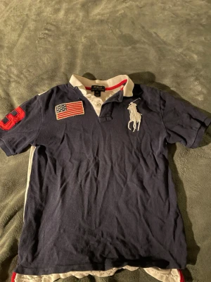 Blå och vit polotopp från Ralph Lauren - Snygg polotopp från Ralph Lauren i blått och vitt med klassisk krage. Framsidan har en stor broderad logga, amerikansk flagga och röd siffra på ärmen. Toppen har korta ärmar och knäppning vid halsen. Perfekt för en sportig och stilren look.