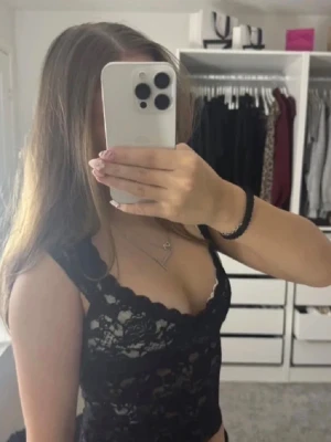 Svart spetsig croppad linnetopp - Säljer en svart croppad linnetopp i spets med v-ringad hals och breda axelband. Toppen har ett genombrutet spetsmönster som ger en snygg och feminin look. Perfekt att styla med jeans eller kjol för en trendig outfit. Den passar både folk med storlek S och XS