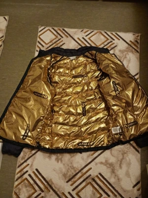 Blå dunjacka med guldigt foder från Columbia - Den blå jackan på bilden är en Columbia dunjacka som använder märkets Omni-Heat värmereflekterande teknologi, vilket syns tydligt på det karaktäristiska guldfärgade fodret inuti.  Produktegenskaper Märke: Columbia Färg: Mörkblå utsida, guldfärgat foder Teknologi: Utrustad med Omni-Heat Reflective Technology, som reflekterar kroppsvärmen för extra värme utan extra vikt. Vissa avancerade versioner, som Omni-Heat Infinity, har ett guldmönstrat foder som beskrivs som guldstandarden inom värmeisoleri