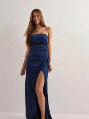 Marinblå axelbandslös långklänning - Säljer en marinblå axelbandslös långklänning i glansigt material från Nelly. Klänningen har en snygg slits framtill och draperad detalj i sidan som ger en elegant look. Klänningen är helt ny, aldrig använd och har prislappen kvar. Perfekt till studentbal eller andra festligheter.  Nypris 799 kr, mitt pris 650 kr  Pris kan diskuteras