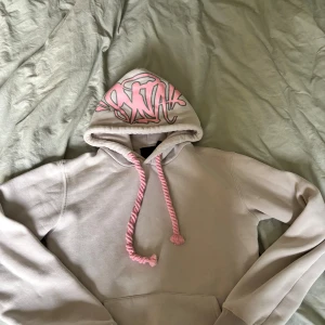 Beige hoodie från Syna world med rosa detaljer - Säljer en beige hoodie från Syna world med rosa snörning och graffiti-inspirerat tryck på huvan. Tröjan är tillverkad i 100% bomull och har en klassisk loose passform med långärmade ärmar. Perfekt för dig som gillar streetwear och unika detaljer.