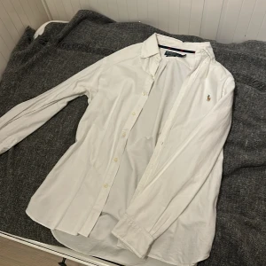 Vit skjorta från Polo Ralph Lauren - Vit skjorta från Polo Ralph Lauren i custom fit med klassisk krage och knappar framtill. Liten broderad logga på bröstet. Skjortan har lång ärm och är gjord i mjuk bomull, perfekt för en clean och stilren look.