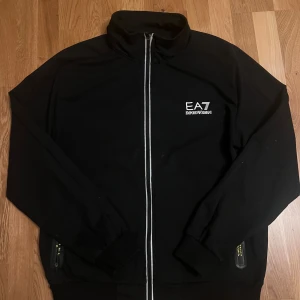 Svart zip upp från EA7 Armani  det är bara L inte 3xl - Svart zip upp från EA7 Emporio Armani med vit logga på bröstet och hel dragkedja framtill. Jackan har högt krage, ribbade muddar och praktiska fickor med dragkedja. Snygg och stilren design som passar till streetwear. Den passar som L den är lite stor för mig bara och jag har M
