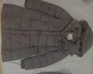 svart  lång pufferjacka med huva - Säljer en svart pufferjacka från lager 157 med huva och långa ärmar. Jackan har quiltad design med rutmönster och knappar framtill. Perfekt för kalla dagar och har en stilren look som funkar till det mesta.