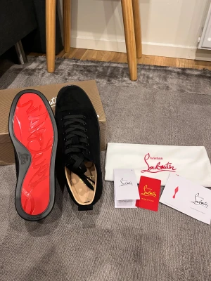 Christian Louboutin svarta sneakers mocka - Svarta sneakers från Christian Louboutin i lyxig mocka med klassisk rund tå och snörning. Sulan är ikoniskt röd med Louboutin-logga, vilket ger en riktigt exklusiv vibe. Insidan är beige och skorna har svarta detaljer och platt sula. Perfekt för dig som gillar stilrena designer.