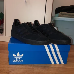 Adidas Gazelle helsvarta sneakers - Säljer ett par helsvarta Adidas Gazelle sneakers med klassiska tre ränder på sidan. Skorna har ovandel i mocka och detaljer i skinn, samt platt sula och snörning. Perfekt för dig som gillar stilrena och tidlösa sneakers med en clean look.