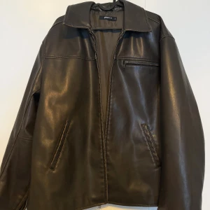 Svart/Brun skinnjacka från Gina Tricot - Snygg skinnjacka från Gina Tricot med klassisk krage, dragkedja framtill och dragkedjor vid ärmsluten. Jackan har raka fickor och en bröstficka med dragkedja. Använd 1 gång. 