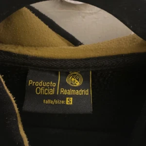 Svart Real Madrid hoodie med dragkedja - Svart hoodie från Real Madrid med gul insida på huvan och broderad logga på bröstet. Stora broderade bokstäver över bröstet, snörning i huvan och praktiska fickor framtill. Dragkedja hela vägen och kontrastsömmar i gult. Perfekt för dig som älskar fotboll och vill ha en sportig look. Från Real Madrids äkta shop