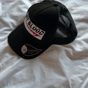 Svart truckerkeps My Blood Atlantis - Svart truckerkeps från Atlantis med böjd skärm och mesh baktill. Framtill finns ett vitt tygmärke med texten 'MY BLOOD' och röd detalj. Justerbar snapback för perfekt passform. Kepsen har en sportig och edgy vibe.