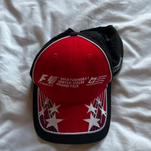 F1 USA Grand Prix keps 2014 - Snygg keps från 2014 Formula 1 United States Grand Prix i Austin, Texas. Kepsen är röd och svart med vita broderade stjärnor och detaljer, samt loggor och banlayout på sidorna. Justerbar baktill och med tydlig racingstil.