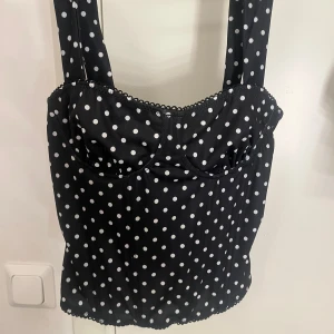 Svart korsett-topp med vita prickar - Säljer en svart korsett-topp med vita prickar och breda axelband. Toppen har markerade kupor, dekorativ spetskant upptill och knäppning med hyskor baktill. Materialet känns mjukt och följsamt, perfekt för dig som gillar retrovibbar och coola detaljer.