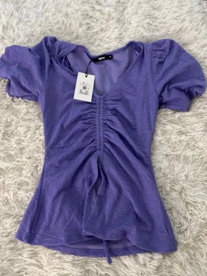 Lila omlottopp med snörning från Bikbok - Supersöt lila topp från Bikbok i storlek XS. Modellen har omlottdetalj och snörning framtill som ger en snygg rynkad effekt. Puffiga korta ärmar och v-ringad hals. Materialet är mjukt och har en lätt struktur som känns skön mot huden.