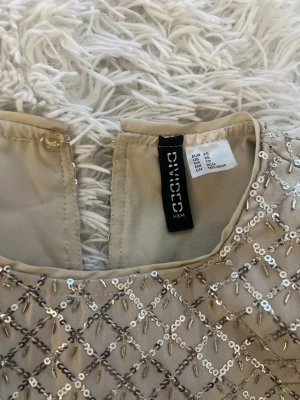 Champagnefärgad glittrig klänning H&M Divided - Champagnefärgad tvådelad klänning från H&M Divided med glittrigt paljettmönster i rutigt. Toppen är ärmlös och croppad, kjolen är kort och rak. Materialet är polyester och har en lyxig satinliknande känsla. Perfekt för dig som vill sticka ut med lite extra bling.