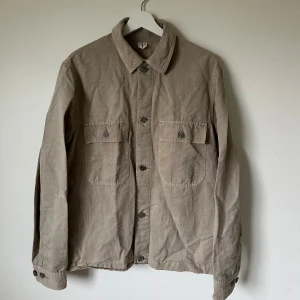Beige overshirt från Arket - Säljer en stilren beige overshirt från Arket i storlek 46. Jackan har två stora bröstfickor med knappar, klassisk krage och knäppning framtill. Tillverkad i mjuk bomull som ger en avslappnad vibe. Perfekt att slänga över en hoodie eller t-shirt. 5J19
