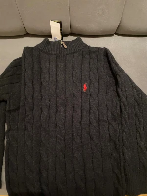Svart stickad polotröja Polo Ralph Lauren - Svart kabelstickad polotröja från Polo Ralph Lauren med röd broderad logga på bröstet. Tröjan har half zip vid halsen och ribbade muddar. Perfekt för dig som gillar klassisk stil med en modern touch.