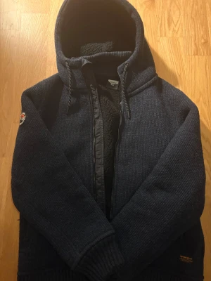 Mörkblå stickad hoodiejacka Superdry - Mörkblå hoodiejacka från Superdry med stickad utsida och mjuk fleece på insidan. Storlek XL på insidan av någon anledning men passar som M bra för 180-185 cm lång. Jackan har huva med snörning, två fickor med dragkedja och Superdry Expedition-märke på ärmen. Perfekt för kalla dagar och chill stil.